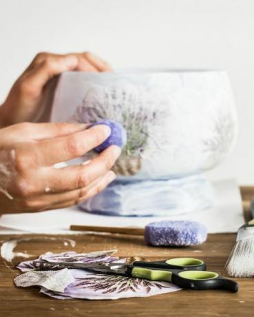 Tijeras de taller de artista de decoupage, esponja, pincel, lápices y manos de pintura de un aficionado decorando un jarrón con un patrón de lavanda