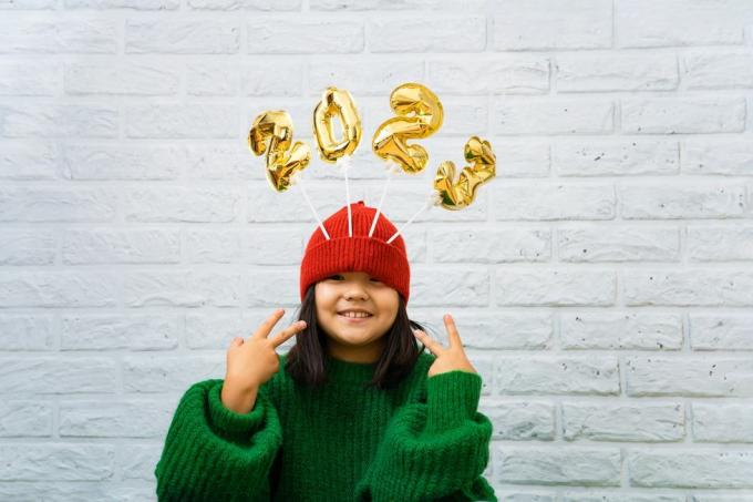 feliz niña asiática con un suéter verde cerca con globos dorados con números 2023 en un espacio de copia de sombrero, fondo de pared de ladrillo blanco, niña mirando la cámara, sonriendo