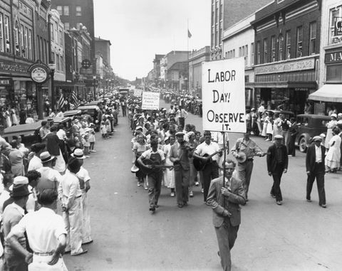 Desfile del día del trabajo en 1934 con gente caminando en la calle mientras sostiene carteles mientras una multitud observa desde las aceras