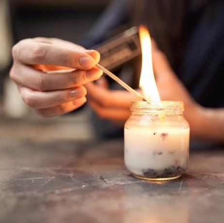 mujer anónima de cosecha con fósforo encendido encendiendo velas aromáticas en un frasco de vidrio colocado sobre una mesa de mármol en casa