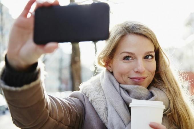 Mujer guapa caminando por las calles de la ciudad en invierno tomándose un selfie y sosteniendo un café para llevar, retroiluminado