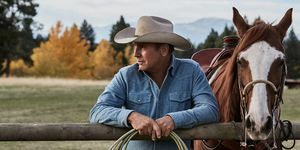 kevin costner en yellowstone junto a un caballo apoyado en una valla con una cuerda en las manos con una camisa de mezclilla azul descolorida y un sombrero de vaquero beige