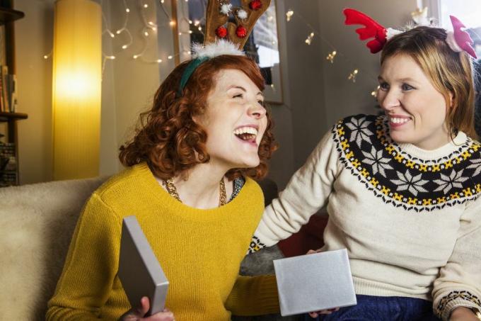 mujer pelirroja sonriendo cuando se sienta en un sofá abriendo un regalo de navidad en una caja plateada hay una mujer con un suéter a su lado sonriendo lleva cuernos rosas y rojos encima de la cabeza