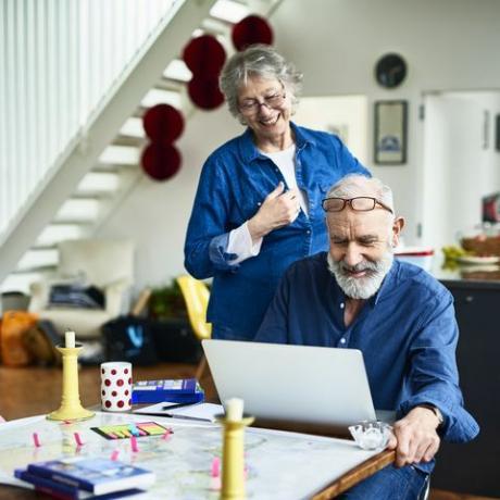 anciana sonriendo y mirando por encima del hombro de su esposo mientras investiga y lee reseñas en línea sobre lugares para quedarse y lugares para visitar, notas adhesivas y guías en la mesa, reserva de vacaciones en línea