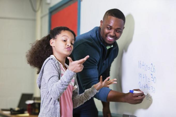 Una niña multirracial de 9 años en clase de matemáticas, parada en la pizarra con su maestra, un joven afroamericano en su 20 años, el maestro mira a su estudiante de primaria, esperando mientras ella intenta encontrar la respuesta, la atención se centra en el chica