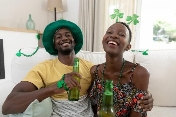 retrato de una pareja afroamericana riéndose con disfraces del día de san patricio sosteniendo botellas de cerveza quedándose en casa aisladas durante el cierre de cuarentena