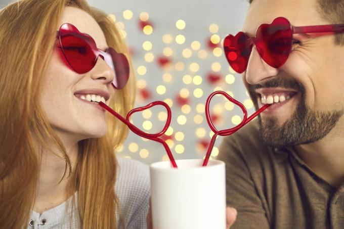 primer plano retrato feliz joven y mujer enamorados con gafas de sol rosas bebiendo de una taza a través de pajitas en forma de corazón, disfrutando de un lindo y divertido momento de pareja en una cita divertida en el día de San Valentín