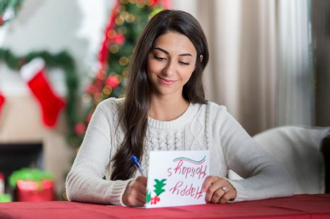 una joven sonriente se sienta en una mesa en su sala de estar decorada y mira hacia abajo concentrada mientras escribe una carta a una amiga para Navidad